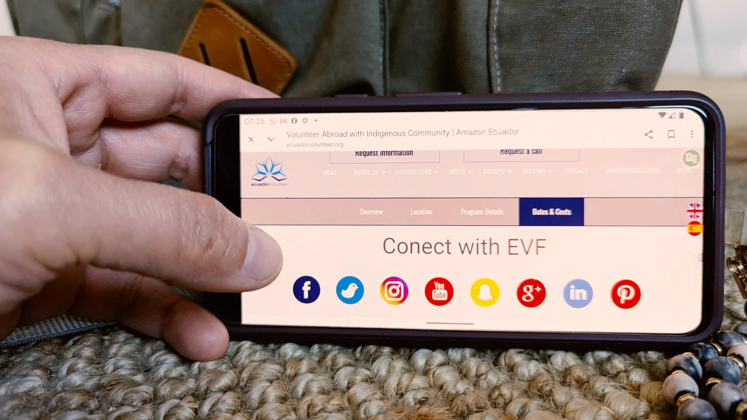 Hand holding a mobile phone with the Ecuador Volunteer website displaying social media icons. Represents communication and contact for volunteers.