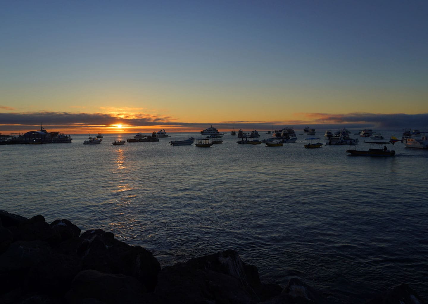 A beautiful orange sunset over the Galapagos harbor, with dozens of boats anchored and the sun reflecting on the calm water.