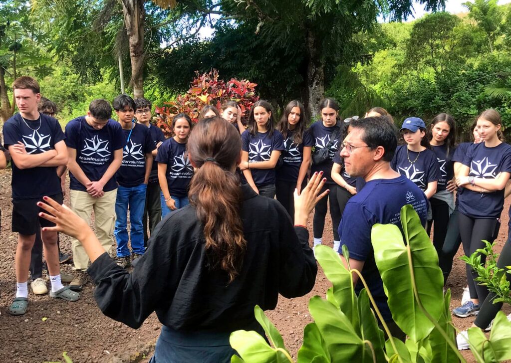 Two leaders from the Galapagos Teen Camp give instructions to a group of teenagers in a natural environment surrounded by vegetation on a sunny day.
