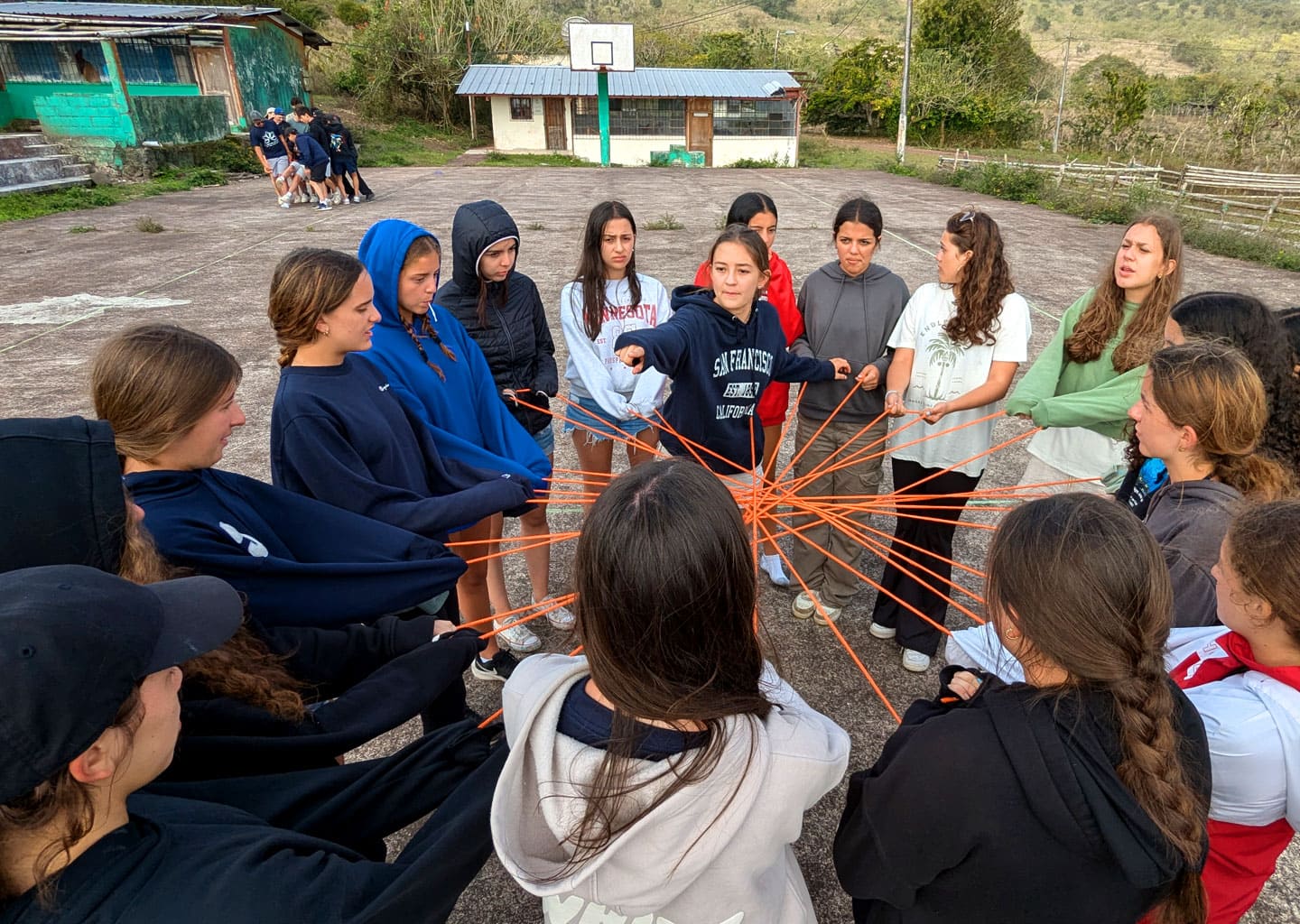 Large group of teenagers participating in a team-building and leadership exercise, holding intertwined orange ropes. Represents the formation of bonds and leadership skills.