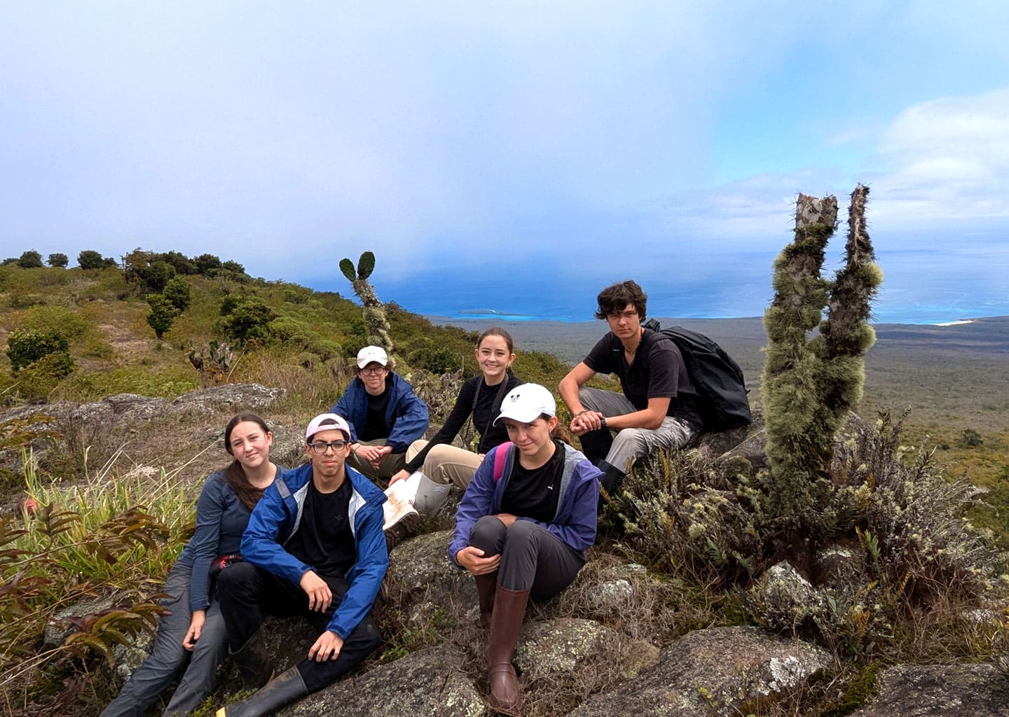Group of five teenagers enjoying a panoramic view of the Galapagos landscape and ocean from the top of a trail, surrounded by volcanic vegetation.
