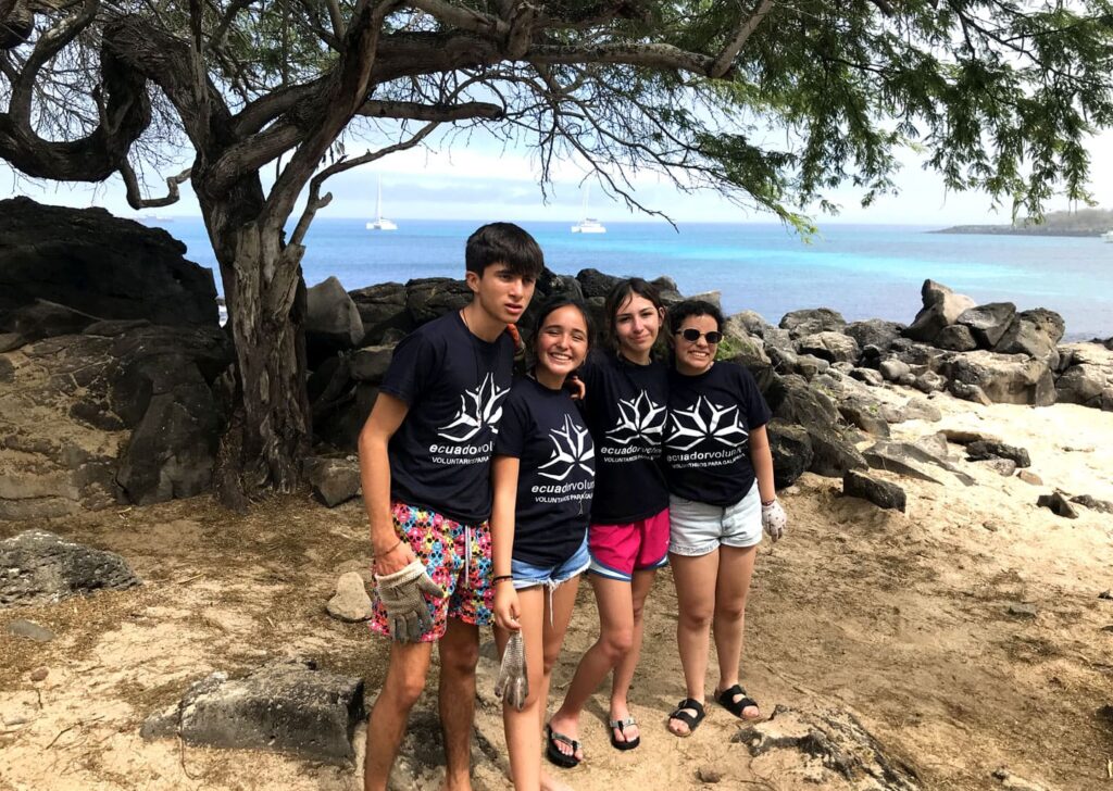 Participants in the Galapagos Teen Camp wearing blue Ecuador Volunteer T-shirts smile and hug each other on a beach while cleaning up trash, with the turquoise sea, sailboats, rocks, and a tree in the background.