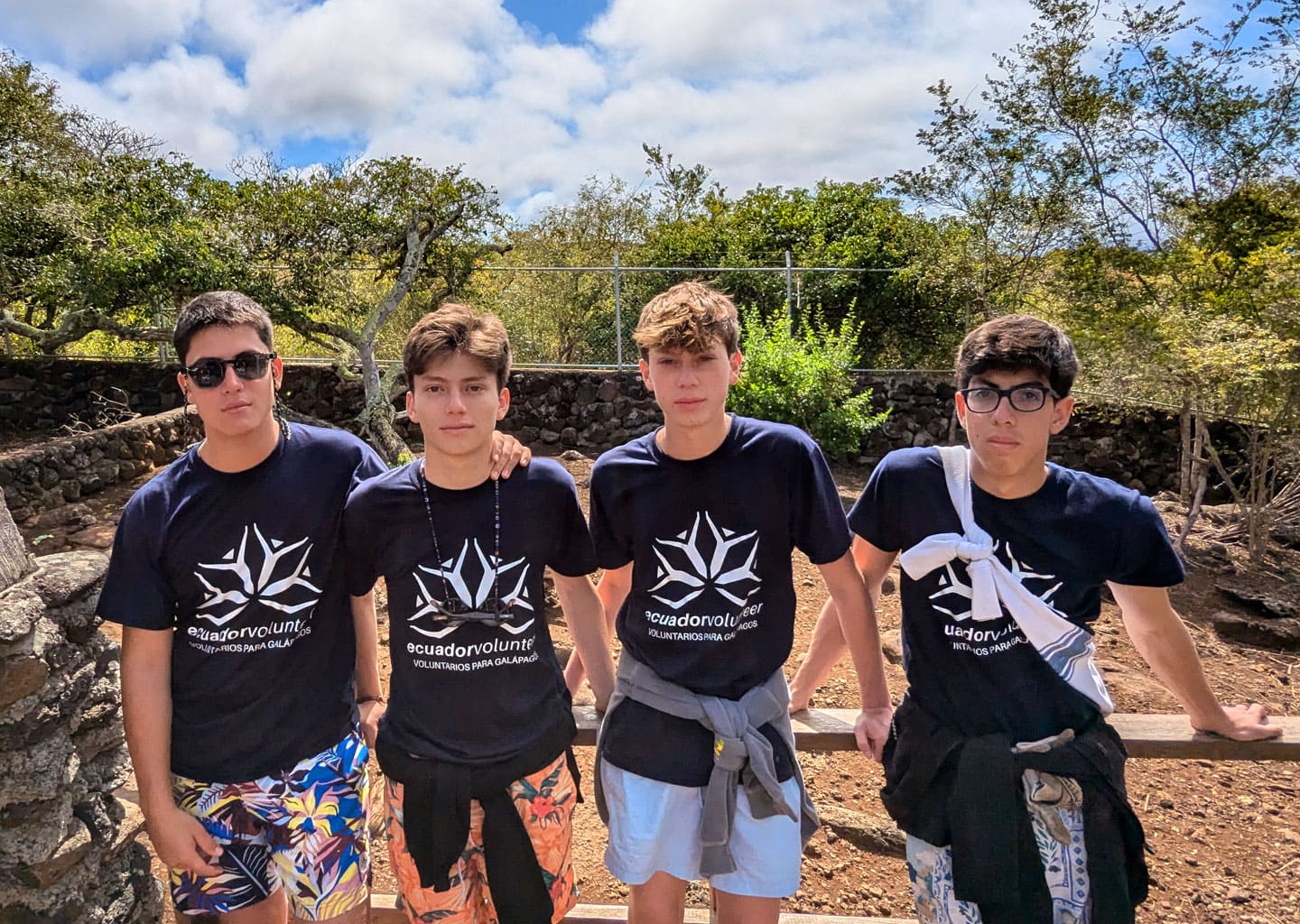 Four teenage volunteers posing on a trail in Galapagos. They wear foundation logo shirts, with endemic vegetation and blue sky in the background.