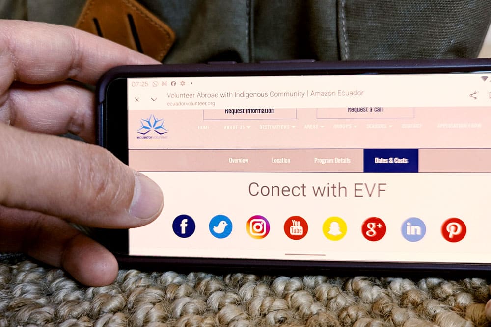 Hand holding a mobile phone with the Ecuador Volunteer website displaying social media icons. Represents communication and contact for volunteers.